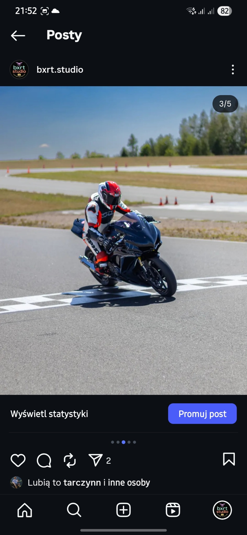 Motocyklista w kombinezonie na czarnym motocyklu sportowym, przejeżdża linię mety na torze wyścigowym w słoneczny dzień. Widoczne opony i kask.