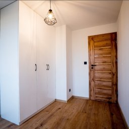 USŁUGI-REMONTOWO-BUDOWLANE-KS-TEAM - Biała zabudowa szafy wnękowej z czarnymi uchwytami, drewniana podłoga i drzwi w nowocześnie wykończonym wnętrzu. Geometryczna lampa wisząca.