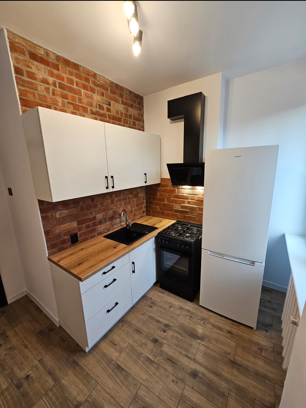 Nowoczesna kuchnia po remoncie: białe szafki z czarnymi uchwytami, ceglana ściana, drewniany blat, czarna kuchenka i okap, biała lodówka, podłoga imitująca drewno.