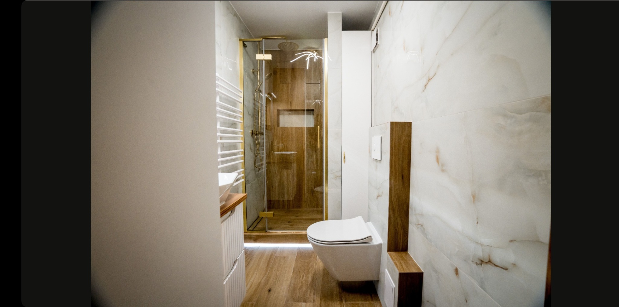 Nowoczesna łazienka z kabiną prysznicową w złotej ramie, marmurowymi płytkami i podwieszaną toaletą. Drewniane akcenty i oświetlenie LED dodają elegancji.