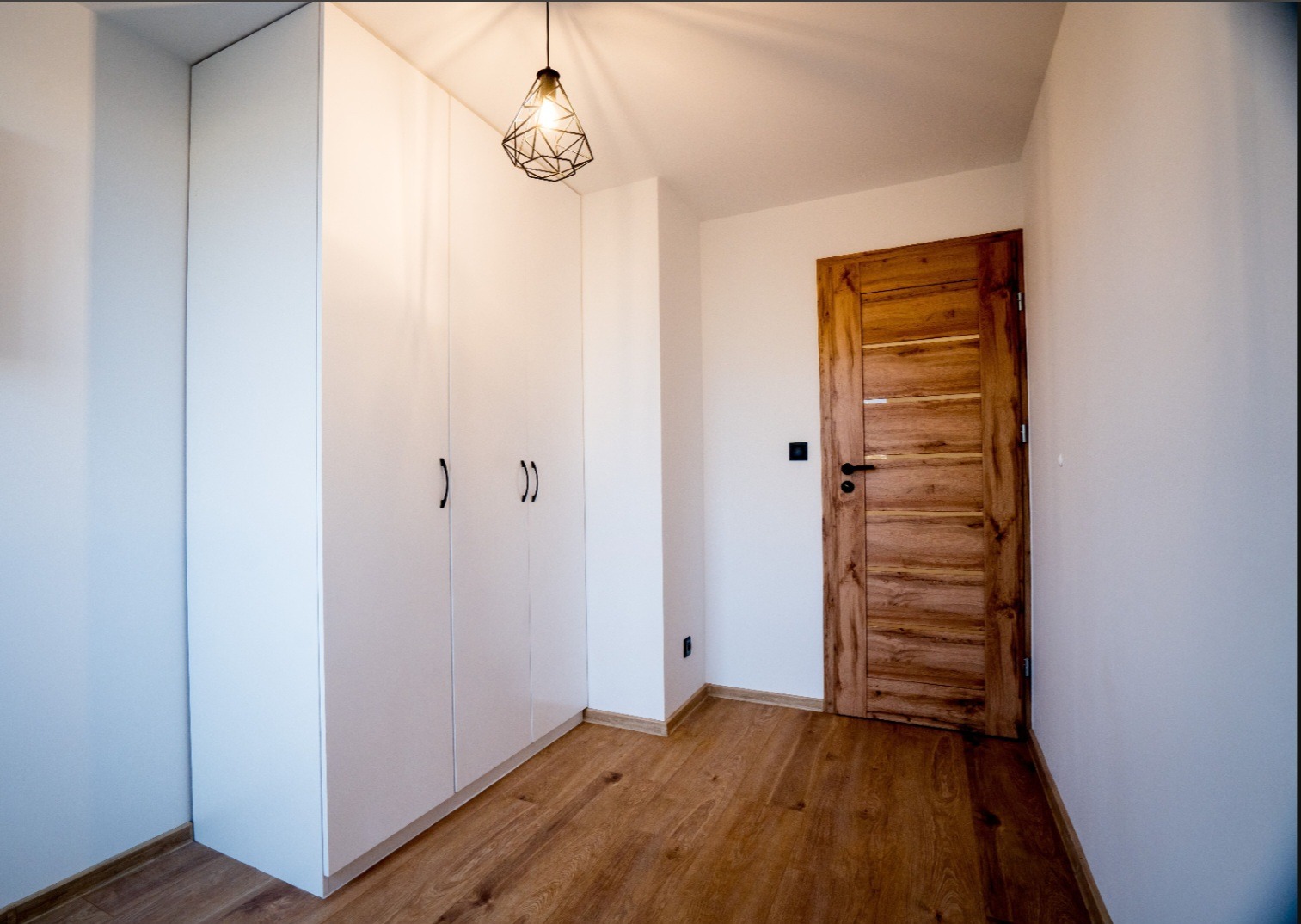 Biała zabudowa szafy wnękowej z czarnymi uchwytami, drewniana podłoga i drzwi w nowocześnie wykończonym wnętrzu. Geometryczna lampa wisząca.