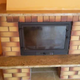 Murowany kominek z cegieł w odcieniach beżu i brązu, z granitowym blatem, widoczna metalowa wkładka kominkowa z szybą.