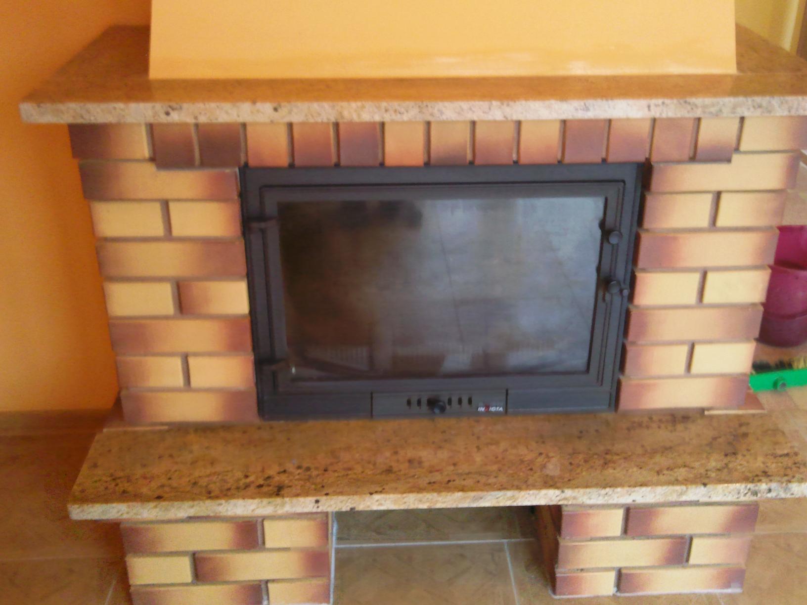 Murowany kominek z cegieł w odcieniach beżu i brązu, z granitowym blatem, widoczna metalowa wkładka kominkowa z szybą.