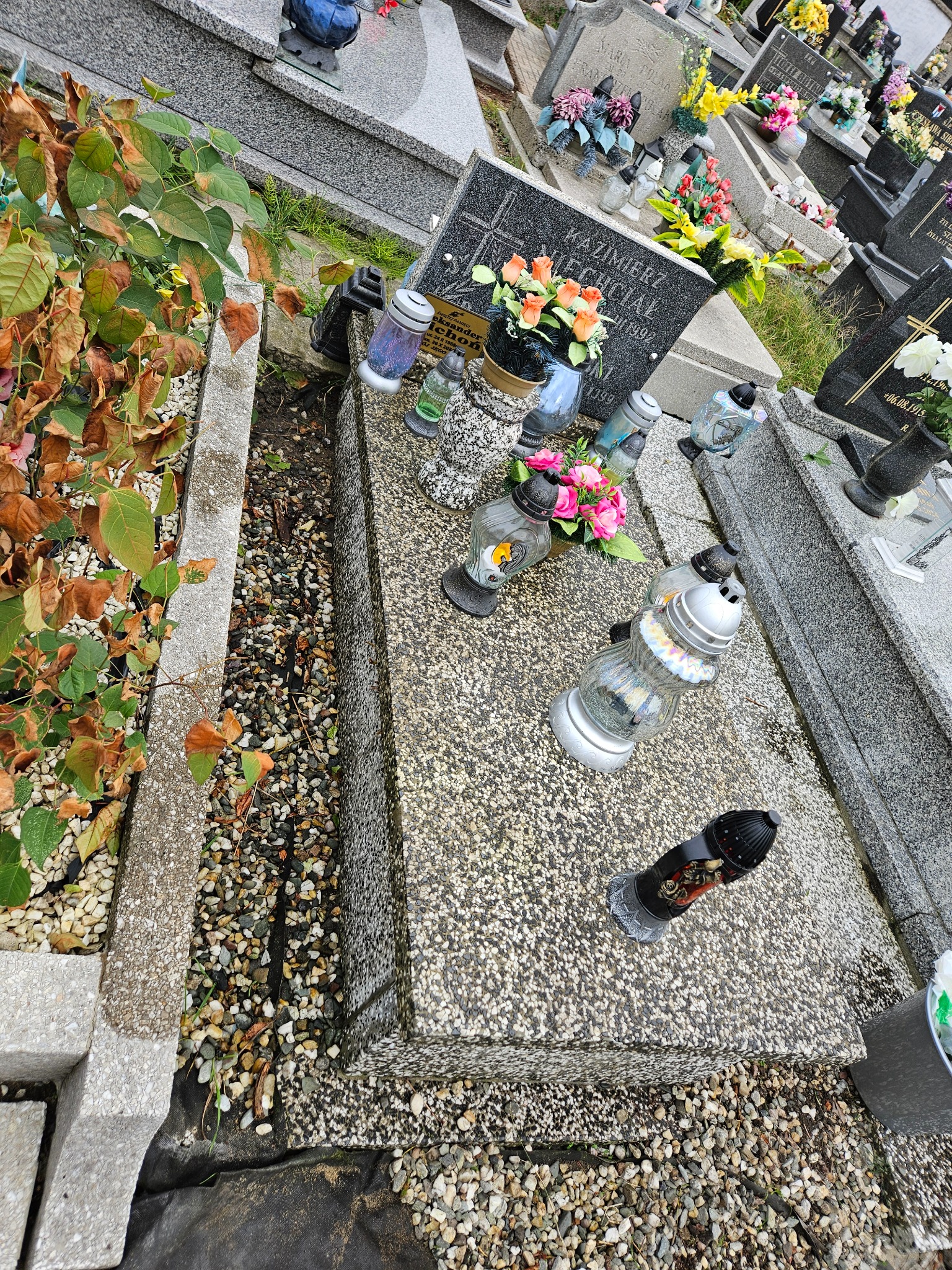 Granitowy nagrobek z kwiatami i zniczami, widok z góry, otoczony innymi nagrobkami na cmentarzu. Jesienne liście w tle.