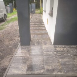 Clean Service &Garden Roman Zalewski - Ułożona kostka brukowa w odcieniach szarości, tworząca ścieżkę do domu. Widoczny fragment elewacji i kolumna w kolorze grafitowym. Po lewej stronie zaparkowany bus.