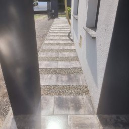 Clean Service &Garden Roman Zalewski - Nowoczesna ścieżka z płyt i żwiru prowadząca wzdłuż białego budynku, widziana zza czarnego słupka. Jasne światło odbija się od mokrej powierzchni.