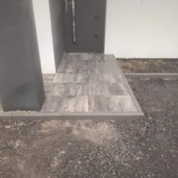 Nowoczesne wejście do domu z szarej kostki brukowej, obrzeża z kostki, widok z góry, grafitowe drzwi i kolumna. Minimalistyczny design.