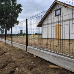 Clean Service &Garden Roman Zalewski - Nowoczesne ogrodzenie panelowe z podmurówką betonową, czarne słupki, widok na budynek w budowie i drzewo. Całość w pochmurny dzień.