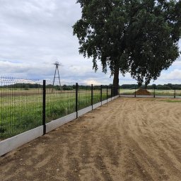 Clean Service &Garden Roman Zalewski - Nowoczesne ogrodzenie panelowe z podmurówką betonową, otoczone piaszczystym terenem i zielenią, z widocznym słupem energetycznym w tle i dużym drzewem po prawej stronie.