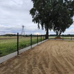 Nowoczesne ogrodzenie panelowe z podmurówką betonową, otoczone piaszczystym terenem i zielenią, z widocznym słupem energetycznym w tle i dużym drzewem po prawej stronie.