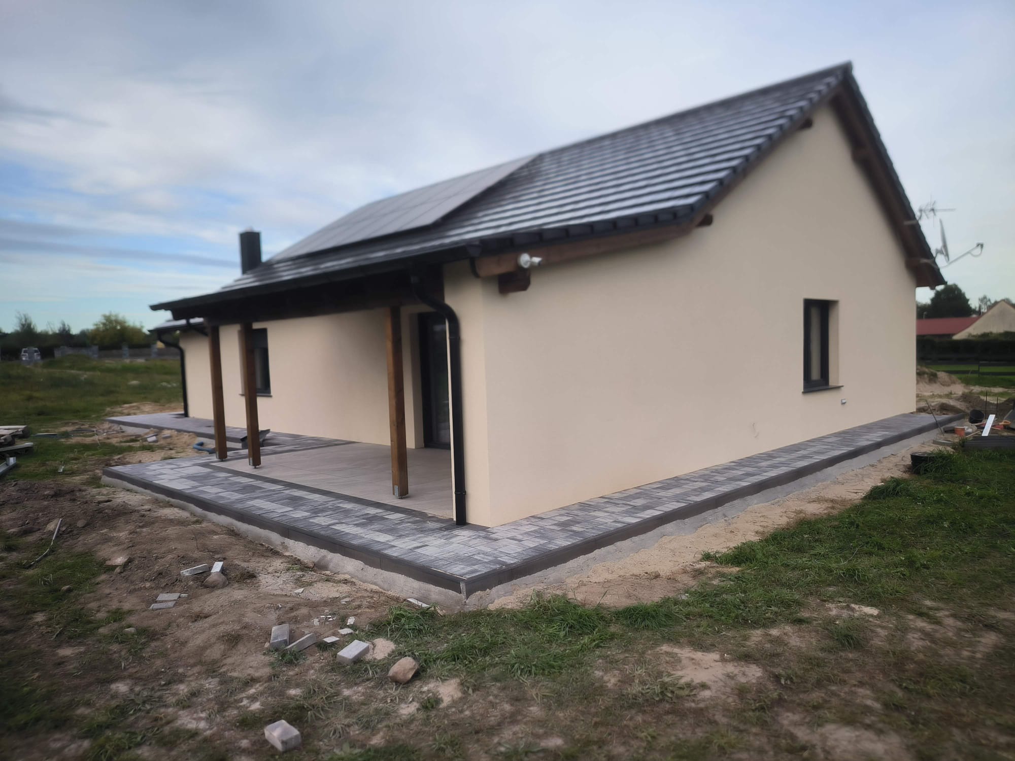 Jasny dom z ciemnym dachem i panelami słonecznymi. Ułożona kostka brukowa wokół budynku. Widoczne niedokończone prace ziemne.