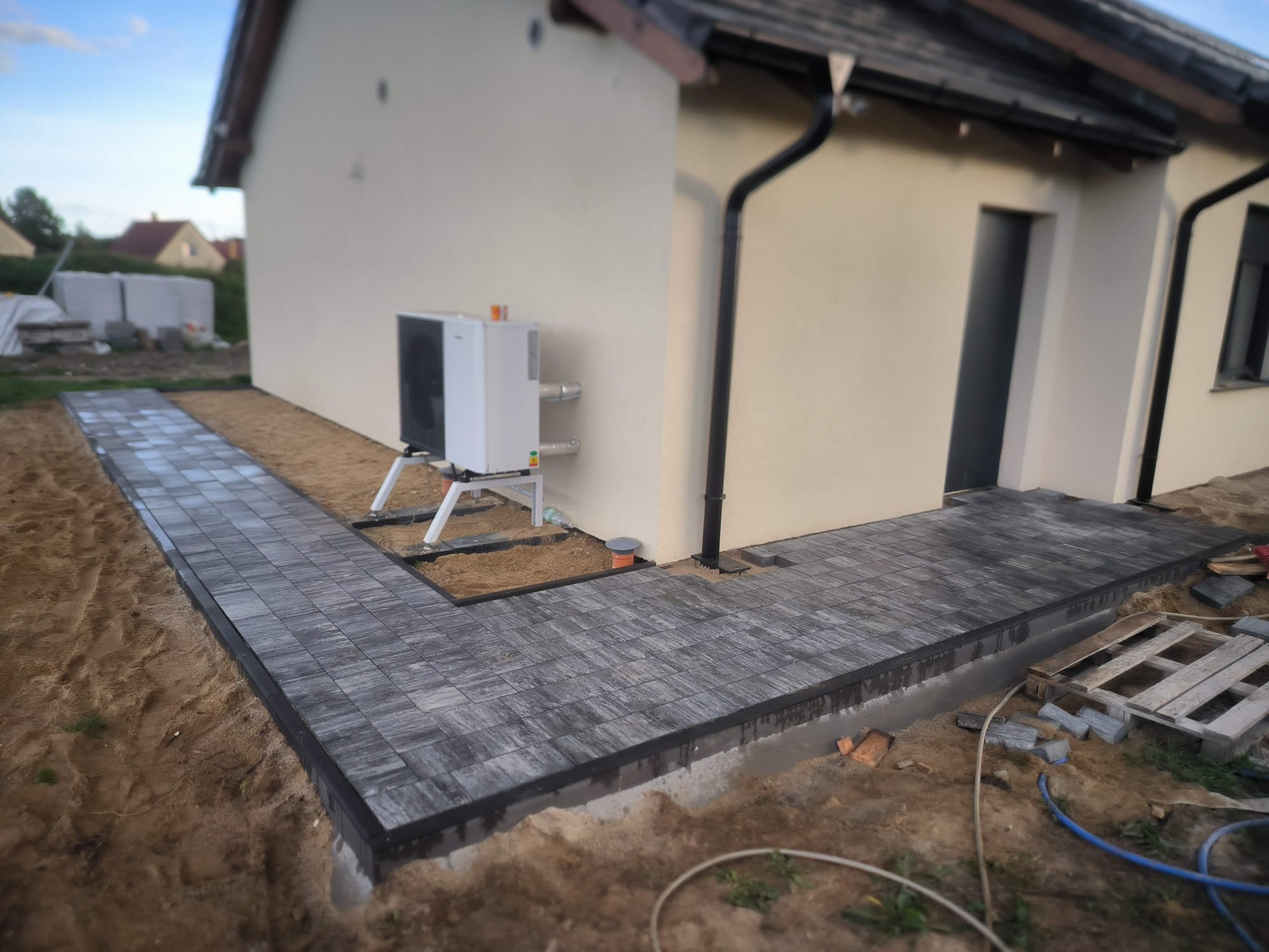 Ułożona kostka brukowa w odcieniach szarości tworzy ścieżkę i taras przy jasnym budynku z pompą ciepła. Widoczne obrzeża i piasek.