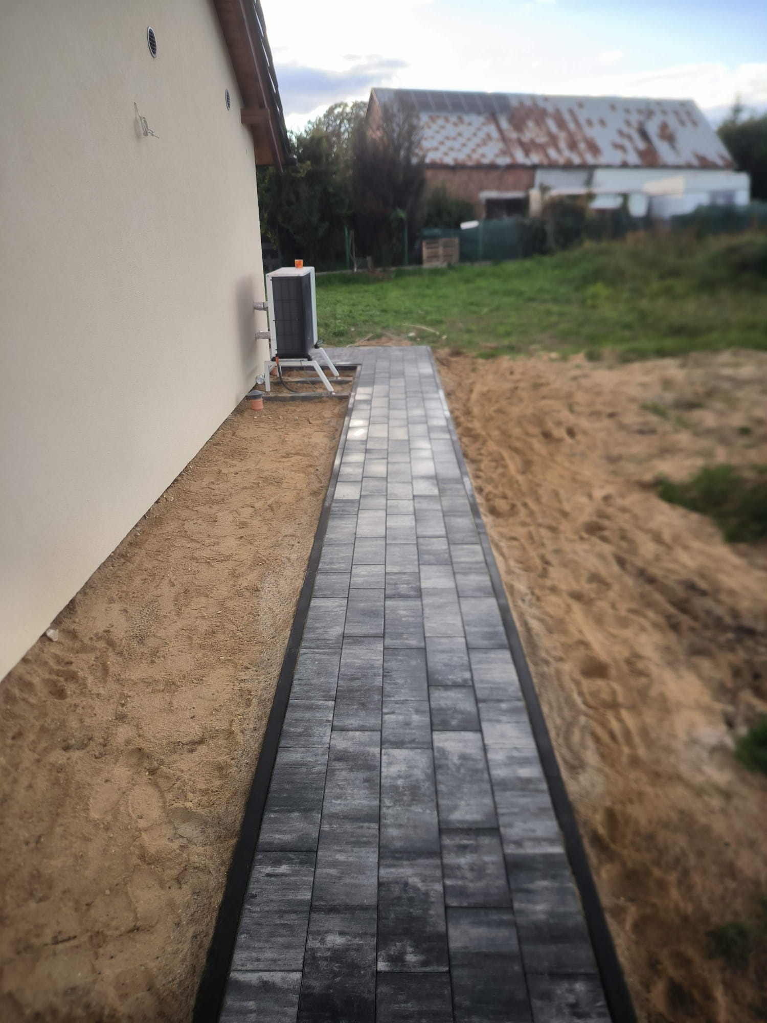 Ułożona ścieżka z szarej kostki brukowej prowadząca wzdłuż budynku z beżową elewacją. Widoczny teren przygotowany pod dalsze prace ziemne.
