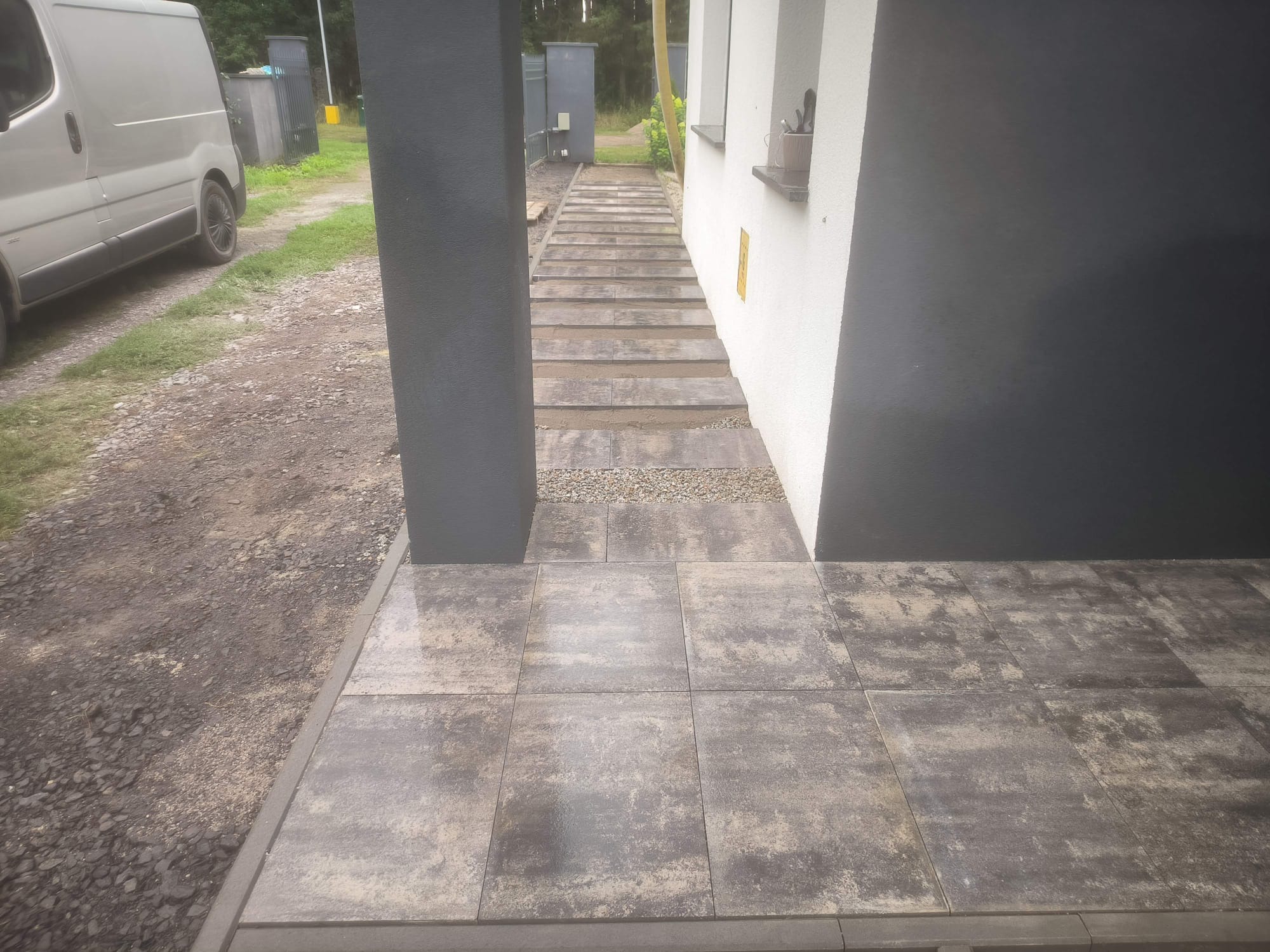 Ułożona kostka brukowa w odcieniach szarości, tworząca ścieżkę do domu. Widoczny fragment elewacji i kolumna w kolorze grafitowym. Po lewej stronie zaparkowany bus.