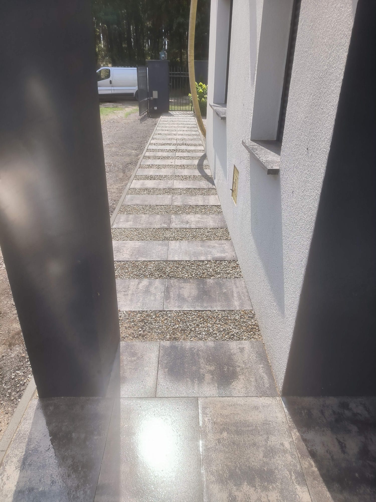 Nowoczesna ścieżka z płyt i żwiru prowadząca wzdłuż białego budynku, widziana zza czarnego słupka. Jasne światło odbija się od mokrej powierzchni.