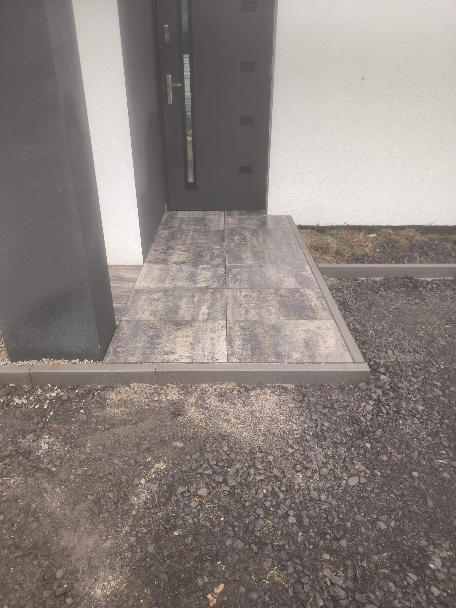 Nowoczesne wejście do domu z szarej kostki brukowej, obrzeża z kostki, widok z góry, grafitowe drzwi i kolumna. Minimalistyczny design.