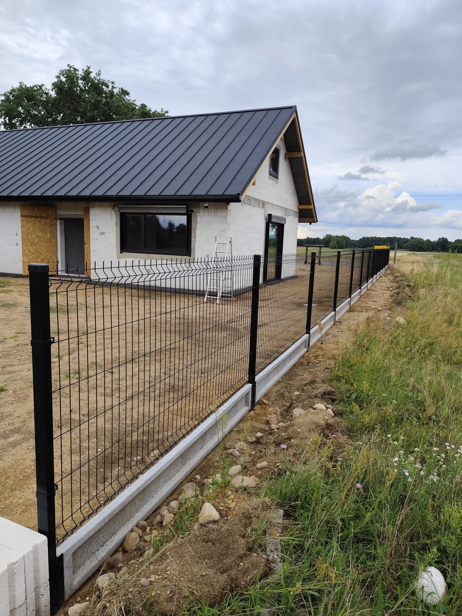 Nowoczesne ogrodzenie panelowe w kolorze czarnym z podmurówką betonową, na tle domu w trakcie budowy i zielonego krajobrazu. Widok z perspektywy.