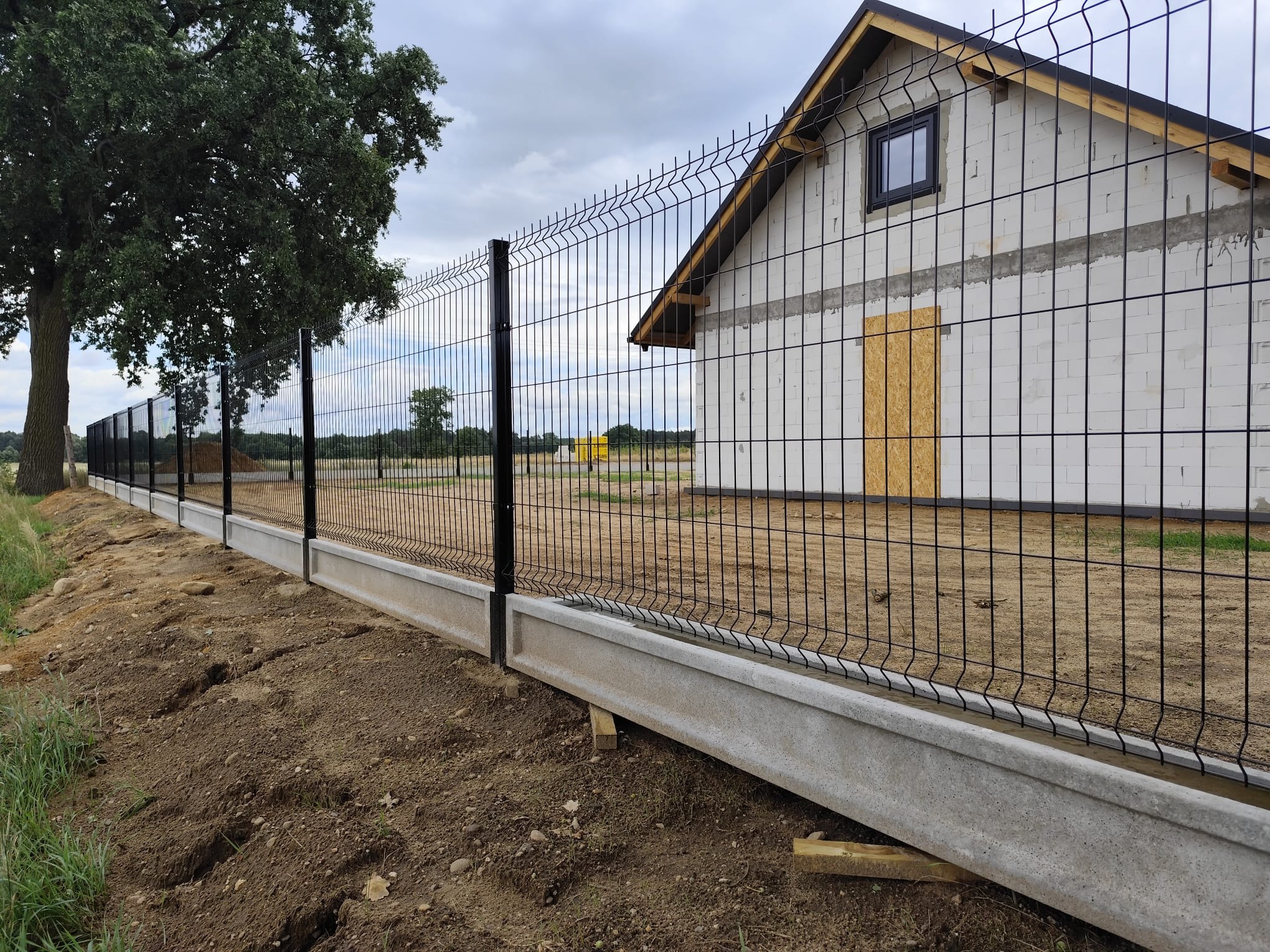 Nowoczesne ogrodzenie panelowe z podmurówką betonową, czarne słupki, widok na budynek w budowie i drzewo. Całość w pochmurny dzień.