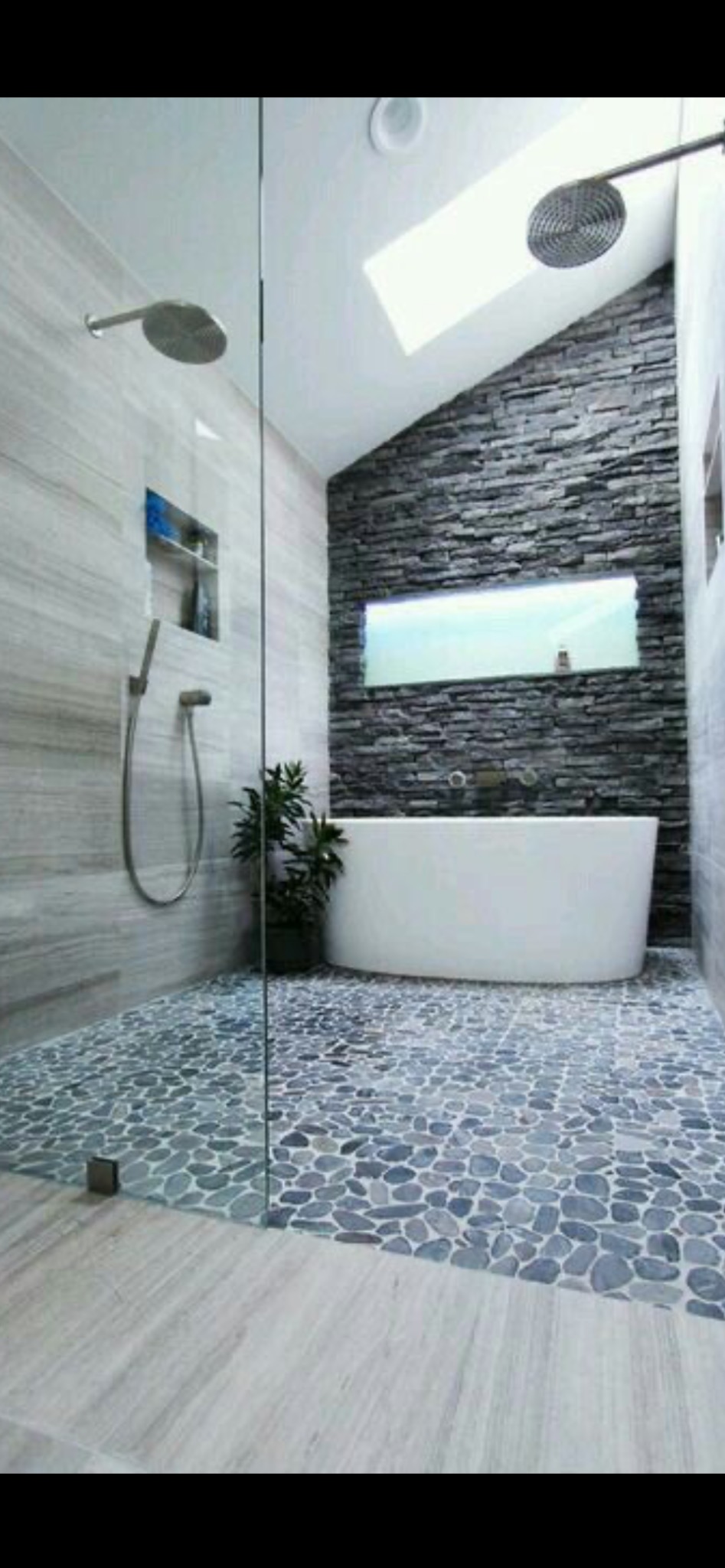 Nowoczesna łazienka z kamienną ścianą, wolnostojącą wanną i podłogą wyłożoną kamieniami. Kabina prysznicowa ze szklaną ścianą. Oświetlenie sufitowe i roślina doniczkowa.