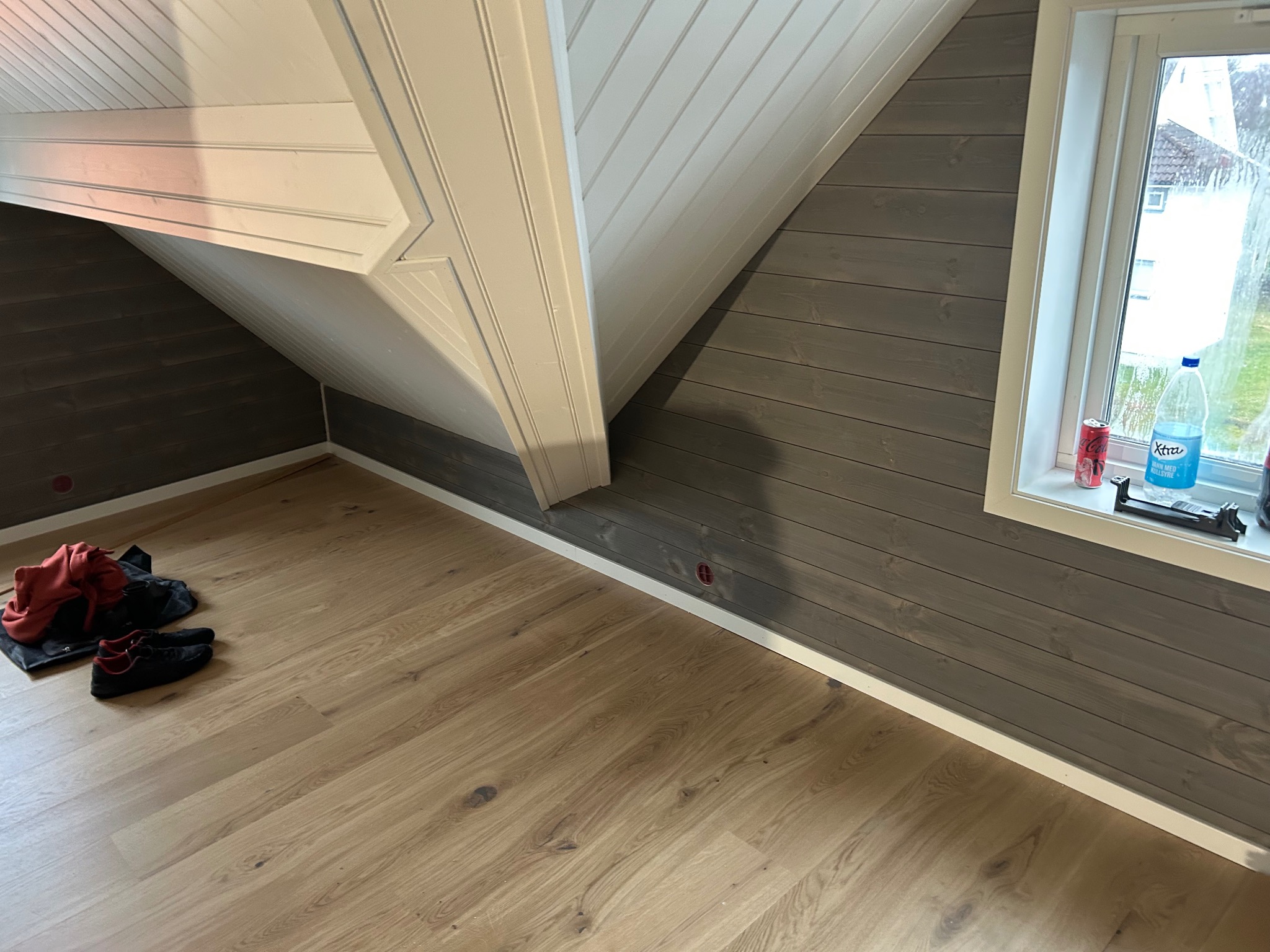 Poddasze po remoncie: panele podłogowe, szare panele ścienne i białe wykończenia. Buty i torba leżą na podłodze. Widok na zewnątrz przez okno.