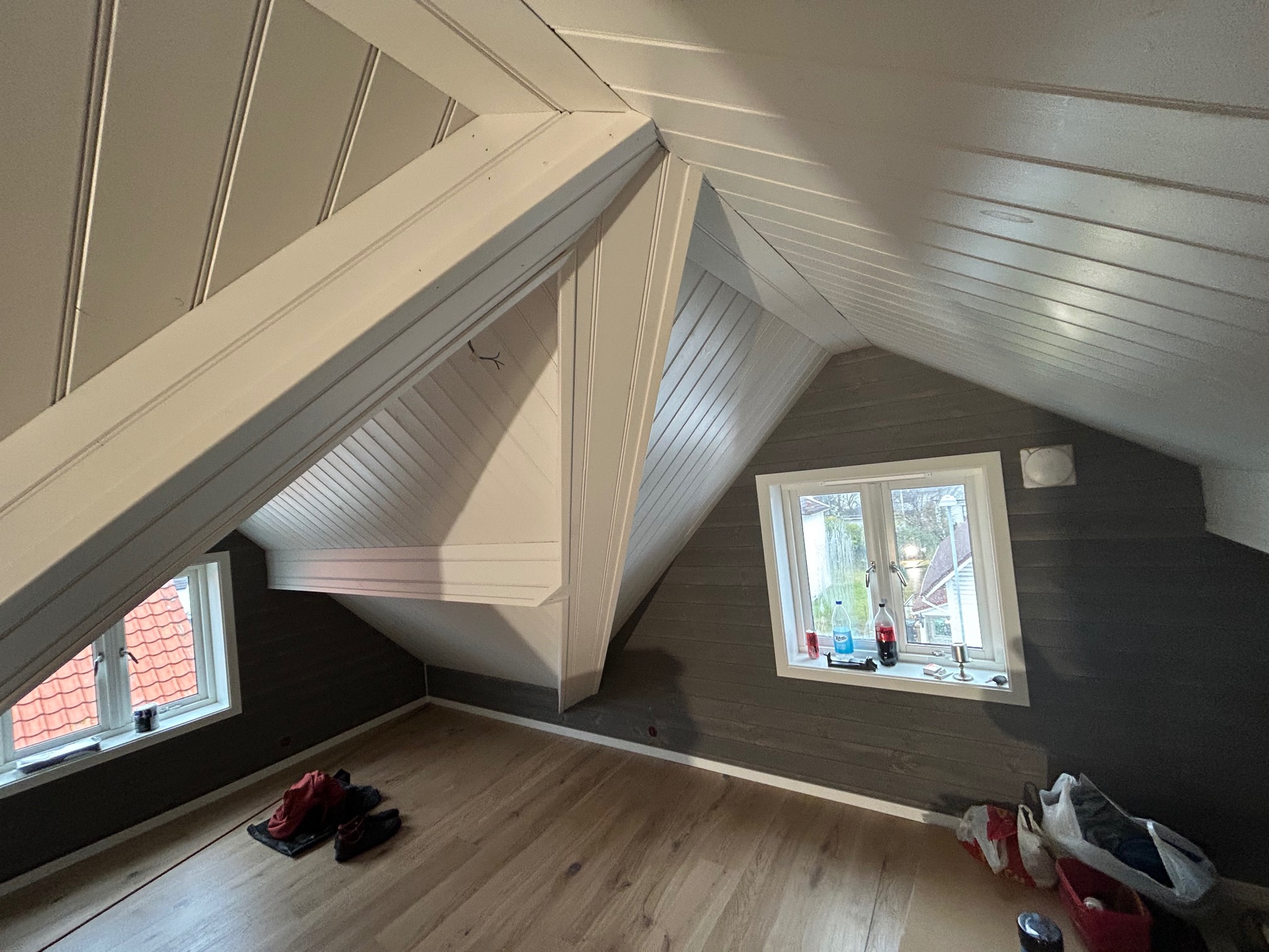 Poddasze po remoncie: drewniana podłoga, szare ściany z boazerią i dwa okna. Widoczne białe belki stropowe i skośny sufit z drewnianą boazerią.