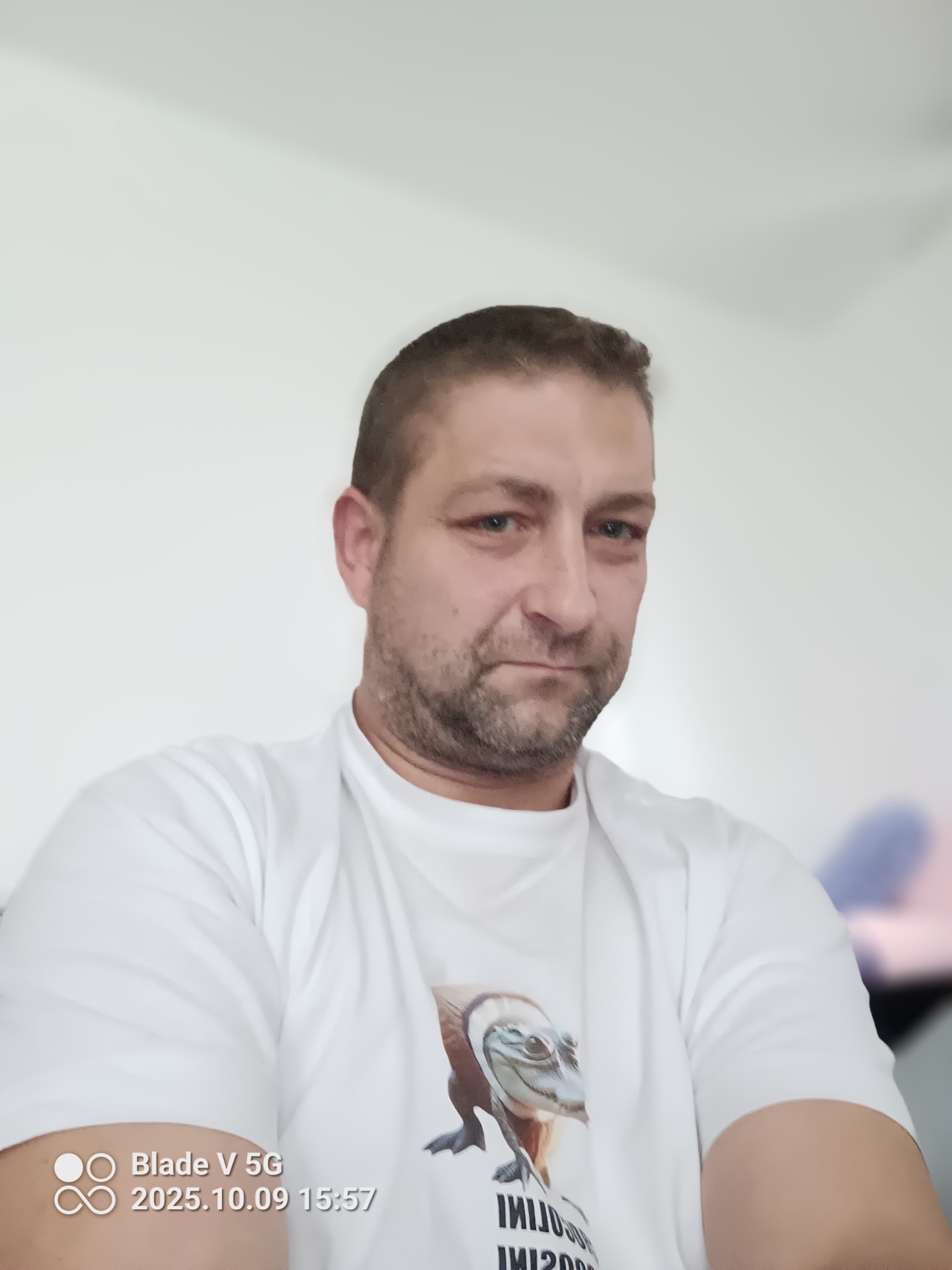 Portret mężczyzny w białej koszulce z nadrukiem, z krótkimi włosami i lekkim zarostem, na rozmytym, jasnym tle.