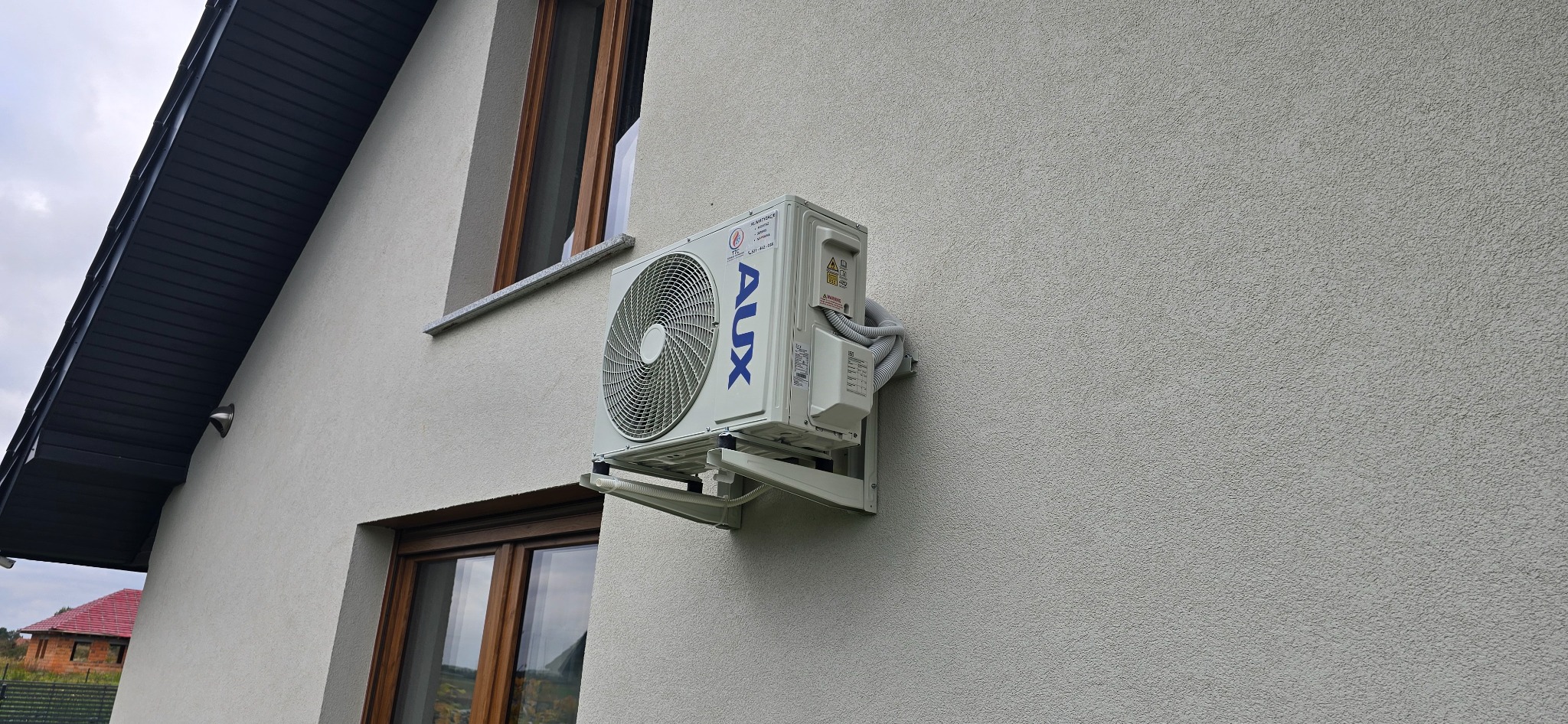 Biała klimatyzacja AUX zamontowana na elewacji domu z widocznymi oknami. Widok z dołu, na tle szarego nieba i fragmentu dachu sąsiedniego budynku.