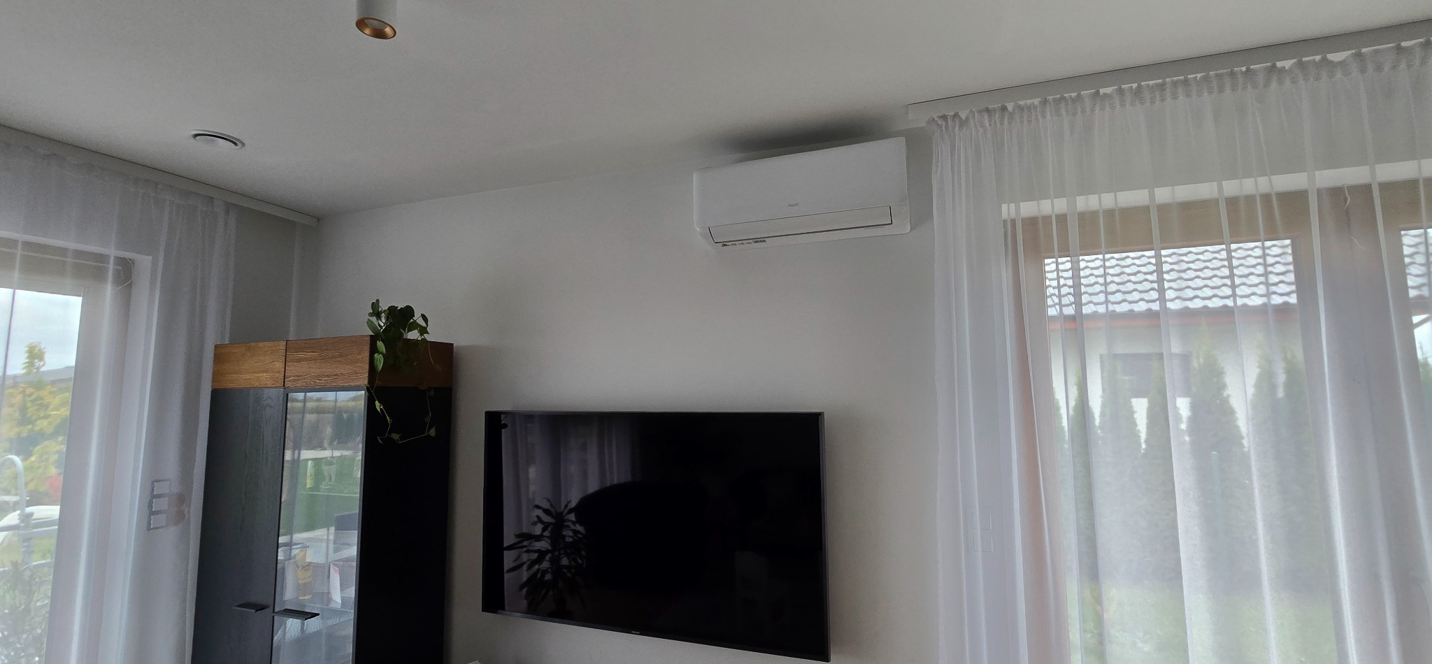 Biały klimatyzator na ścianie w jasnym pokoju z telewizorem i firankami. Widoczny fragment okna i szafki. Minimalistyczny design wnętrza.