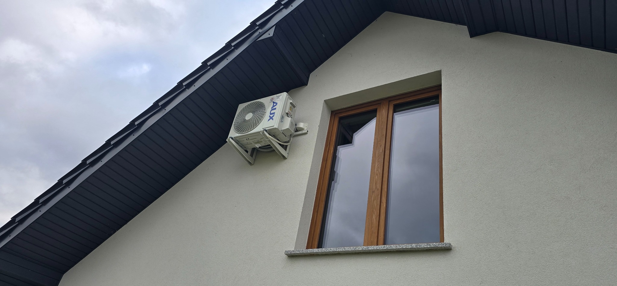 Biały klimatyzator AUX zamontowany na elewacji domu obok okna z brązową ramą. Czarny dach i pochmurne niebo w tle.