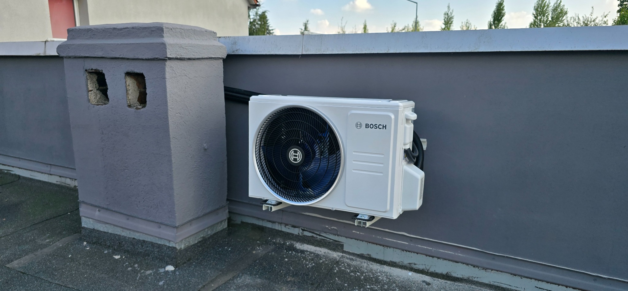 Zamontowana klimatyzacja Bosch na szarym dachu budynku w Kunicach. Widoczny komin i fragment ściany. Czarna rura odprowadzająca skropliny.