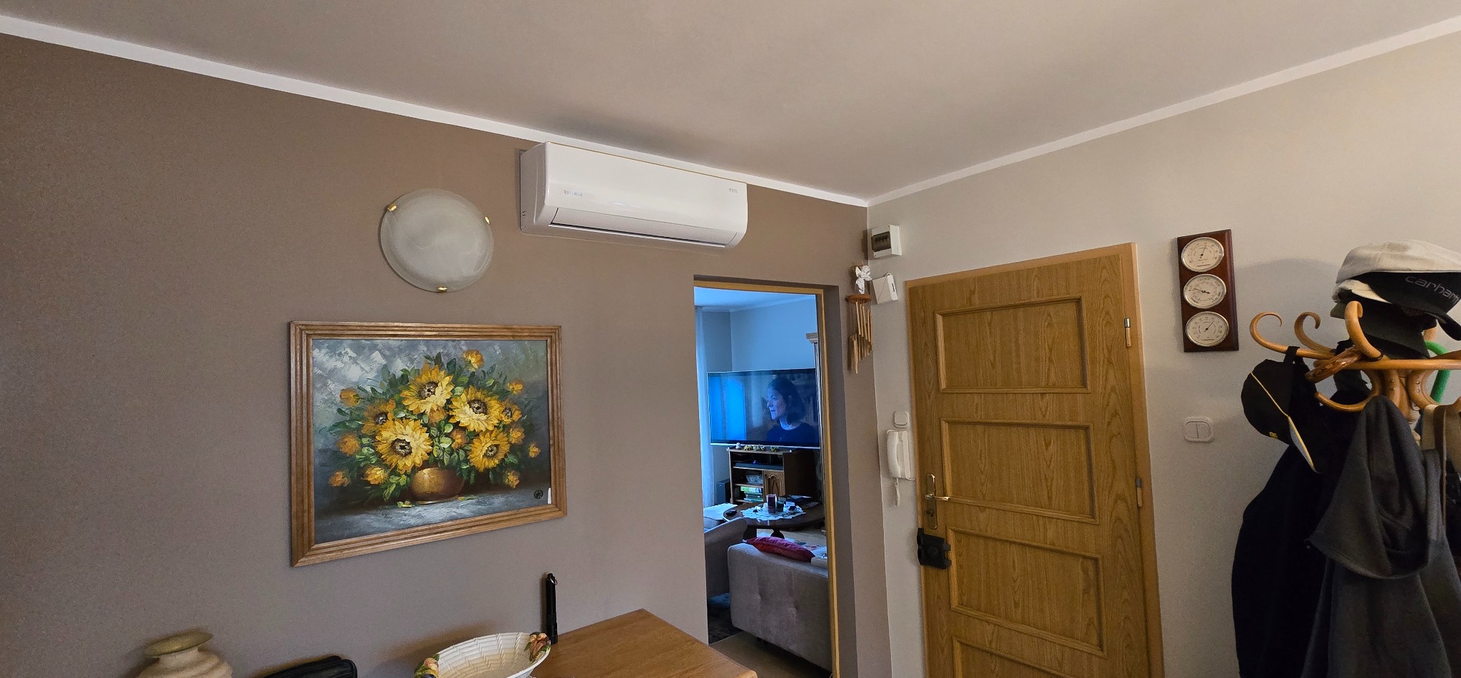 Biała klimatyzacja zamontowana na ścianie w salonie nad obrazem z motywem słoneczników. Widok na wnętrze pokoju przez otwarte drzwi.