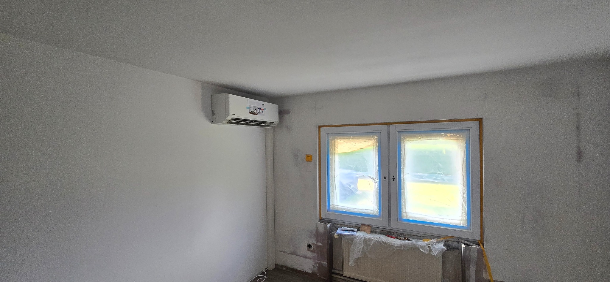 Biała klimatyzacja Panasonic zamontowana na ścianie w pokoju w trakcie remontu; okno zabezpieczone folią malarską. Widoczny grzejnik.
