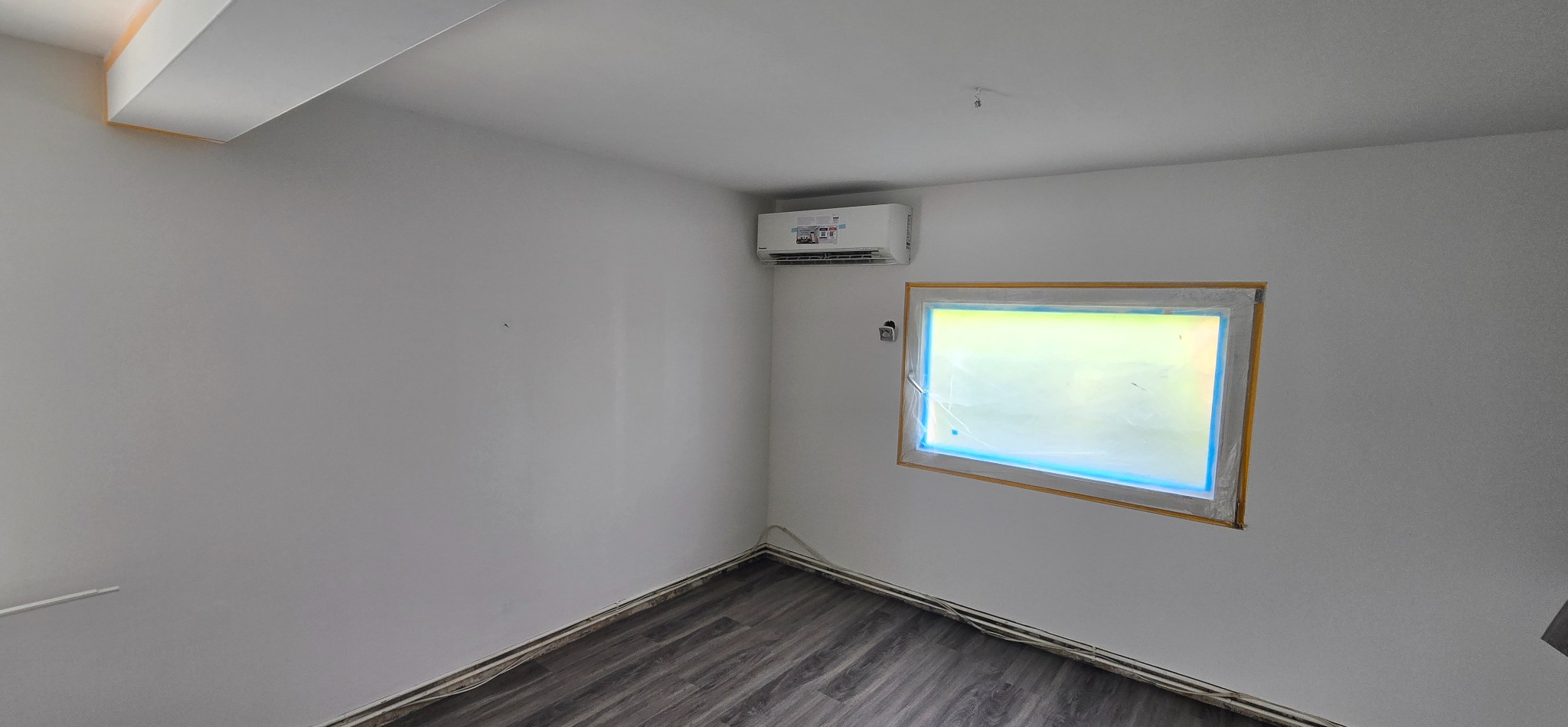 Wnętrze pokoju z zainstalowaną klimatyzacją ścienną. Okno zabezpieczone folią malarską. Podłoga z paneli imitujących drewno. Kabel wystający ze ściany.