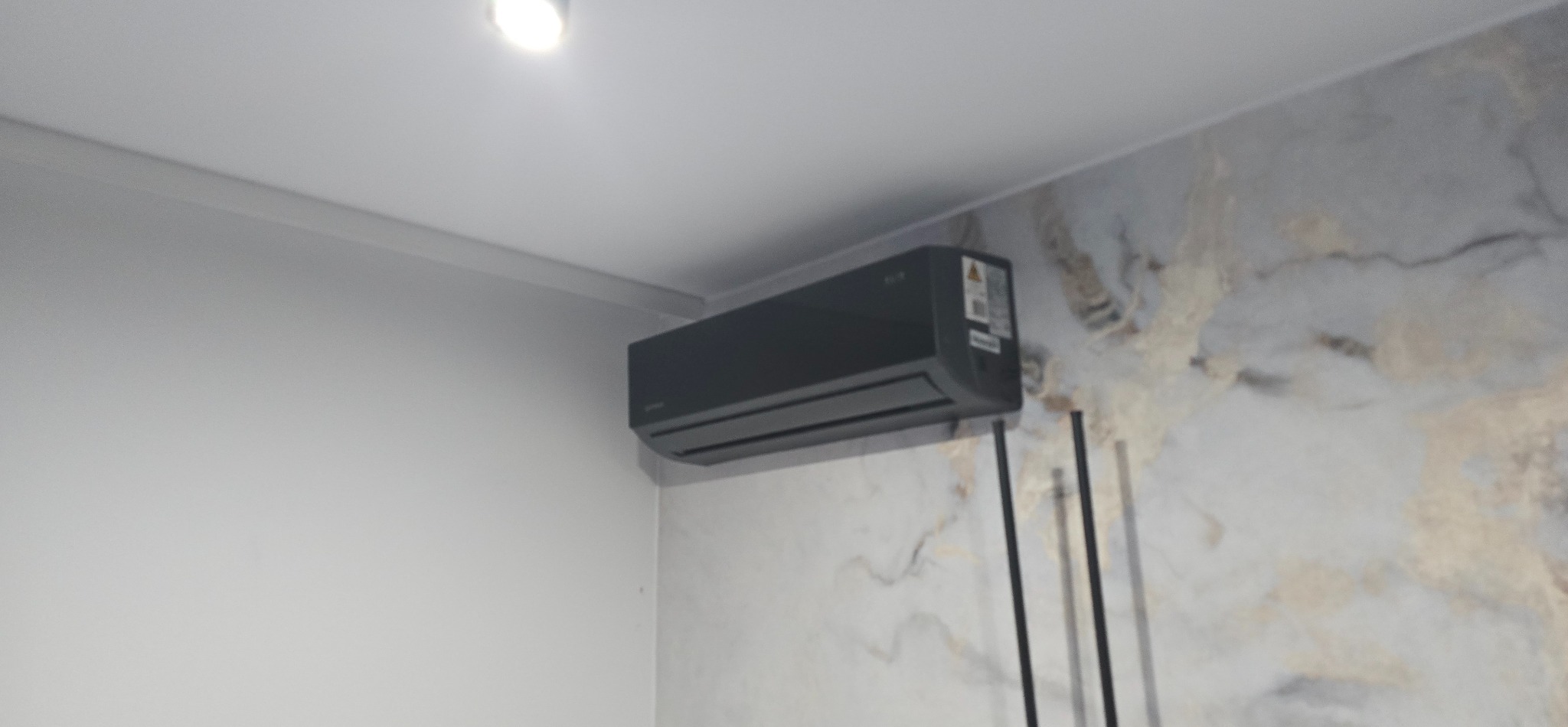 Czarny klimatyzator ścienny na tle marmurowej ściany w jasnym wnętrzu. Widoczne pionowe rurki instalacyjne i sufit z oświetleniem punktowym.