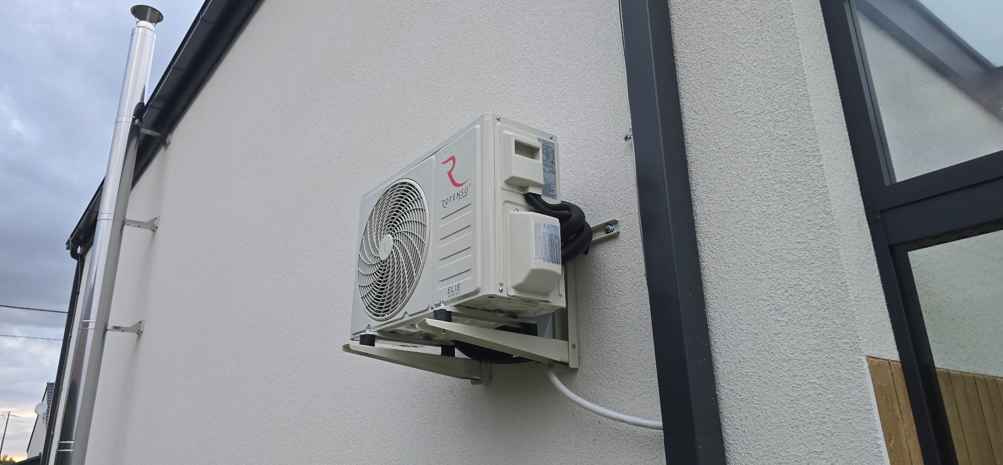 Biała jednostka zewnętrzna klimatyzacji Rotenso, zamontowana na elewacji domu z widocznym kominem i fragmentem okna. Świeży montaż, czyste otoczenie.