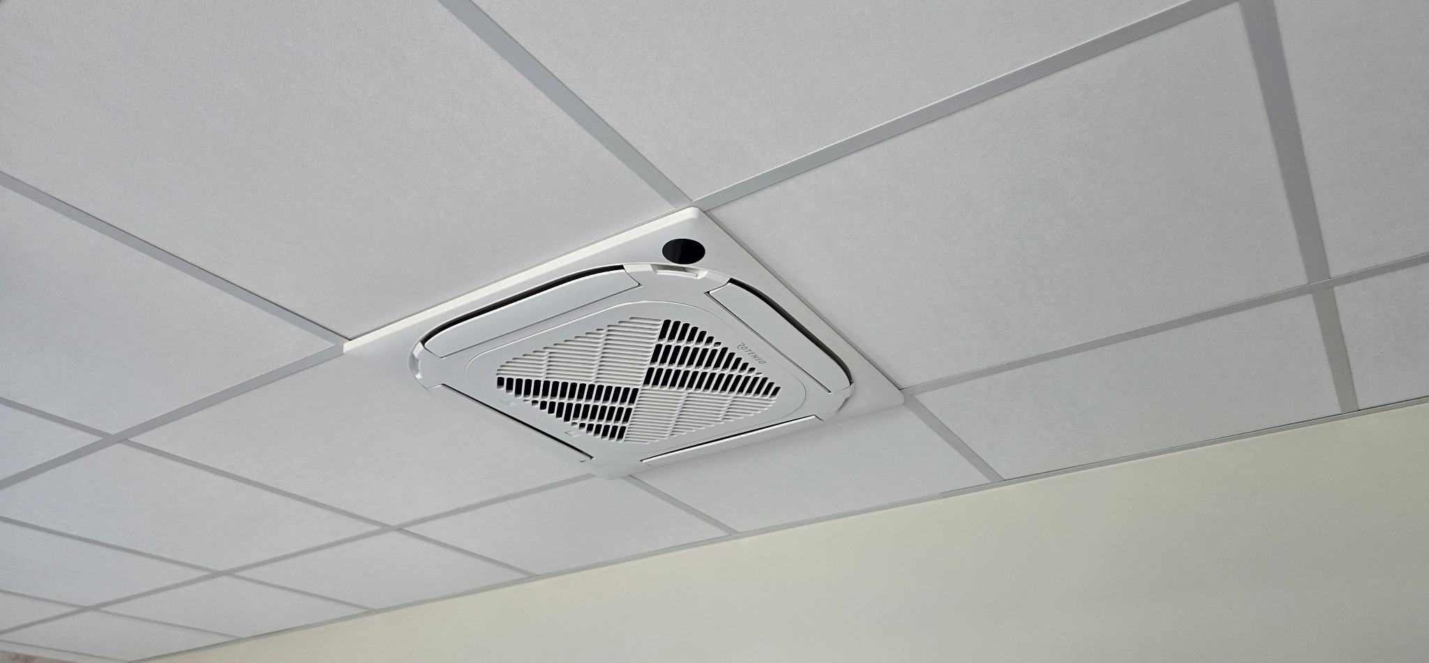 Kasetonowa klimatyzacja sufitowa marki Rotenso, zamontowana w biurze. Widok z dołu, kratka wentylacyjna i czujnik.