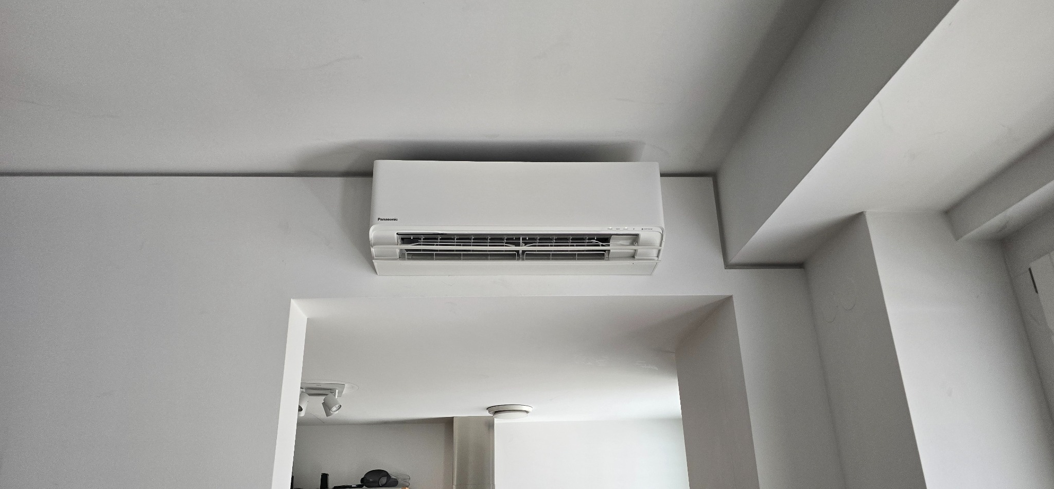 Biały klimatyzator Panasonic zamontowany na ścianie w jasnym wnętrzu, widoczny z dołu, z fragmentem sufitu i przejścia do innego pomieszczenia.