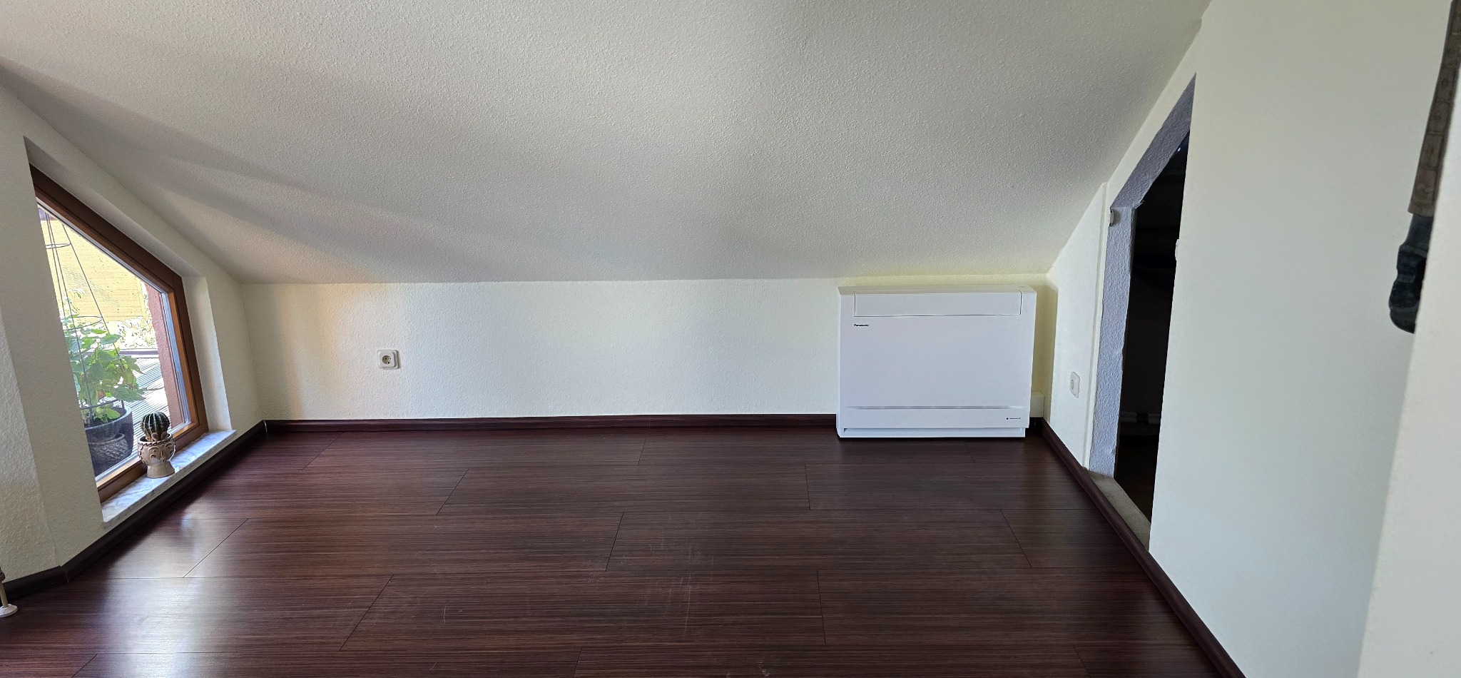 Biała klimatyzacja podłogowa Panasonic w jasnym pokoju na poddaszu z oknem i ciemną podłogą. Widoczny fragment ściany z gniazdkiem elektrycznym.