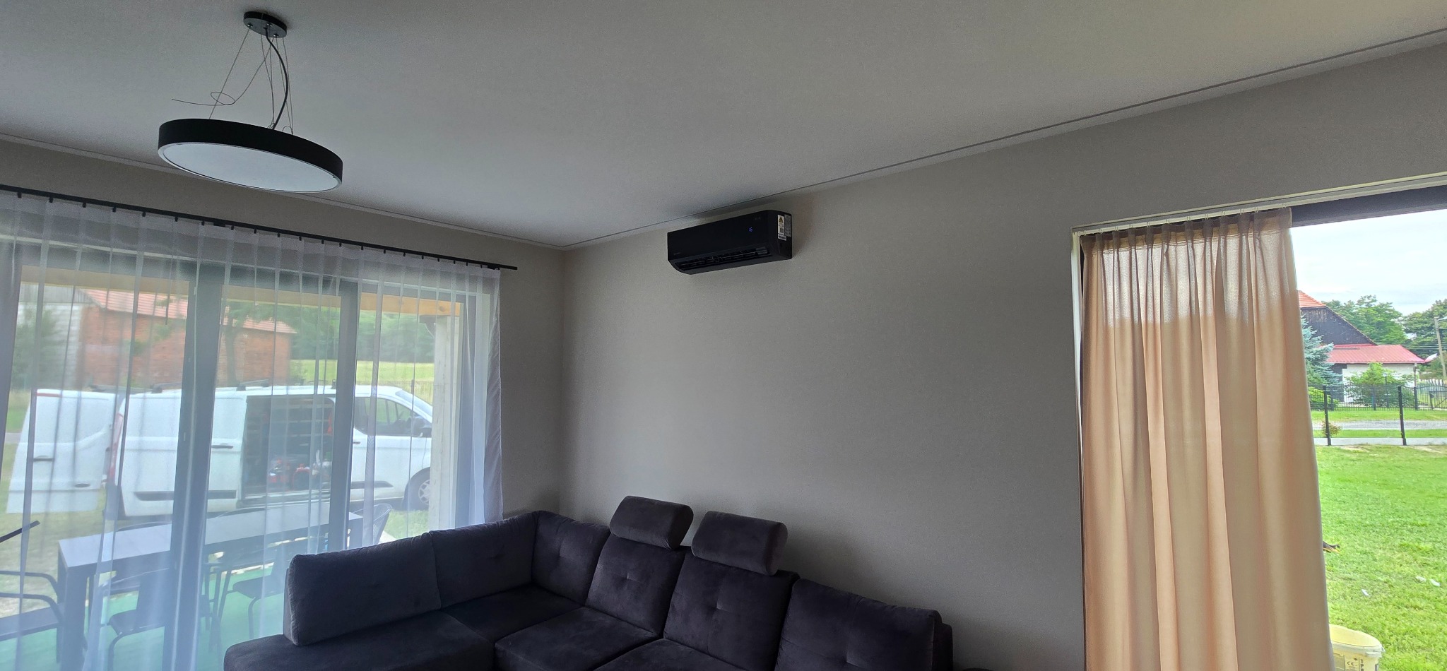 Czarny klimatyzator na ścianie w salonie z szarym narożnikiem. Przez okno widoczny samochód serwisowy. Nowoczesne oświetlenie sufitowe.