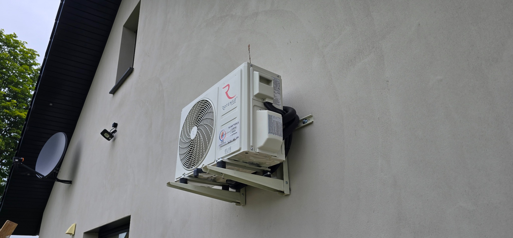 Zamontowany klimatyzator Rotenso Elis Series na elewacji domu. Widoczne przewody i uchwyty montażowe. W tle fragment dachu, okna i antena satelitarna.