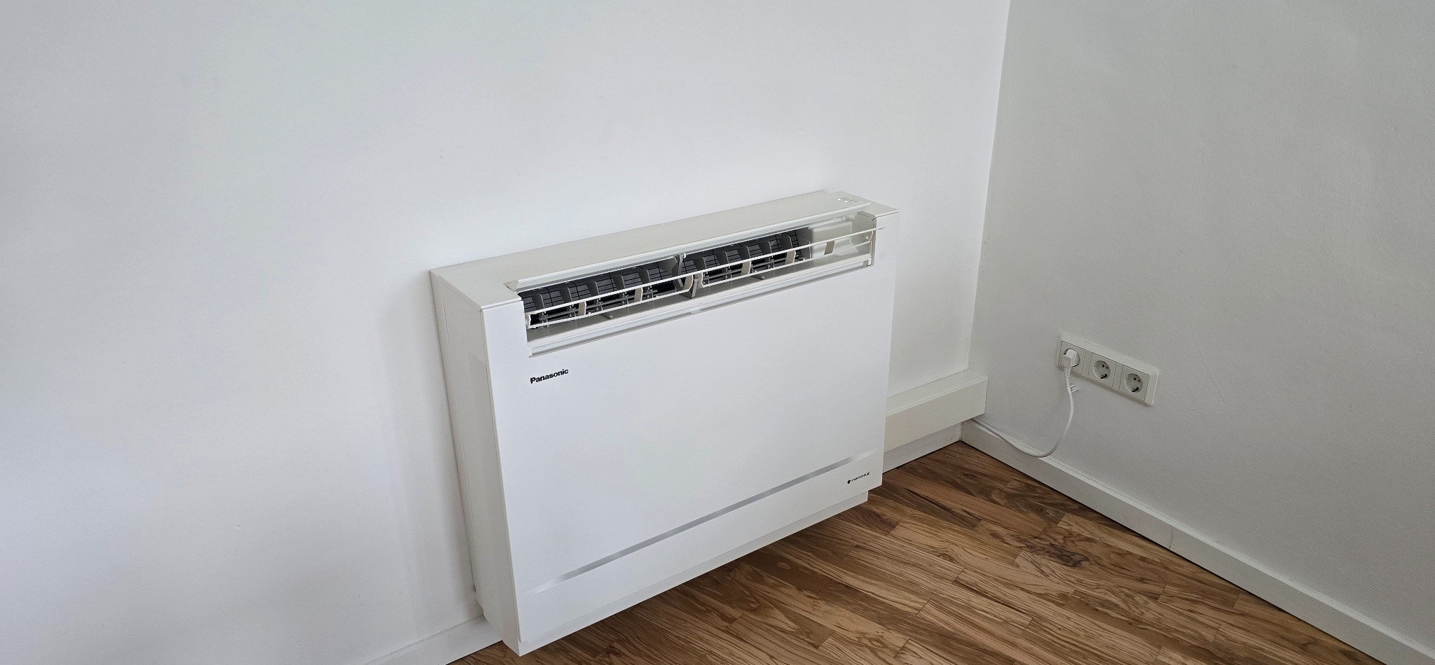 Biała, podłogowa klimatyzacja Panasonic w rogu pomieszczenia z drewnianą podłogą i gniazdkiem elektrycznym na ścianie. Minimalistyczny design.