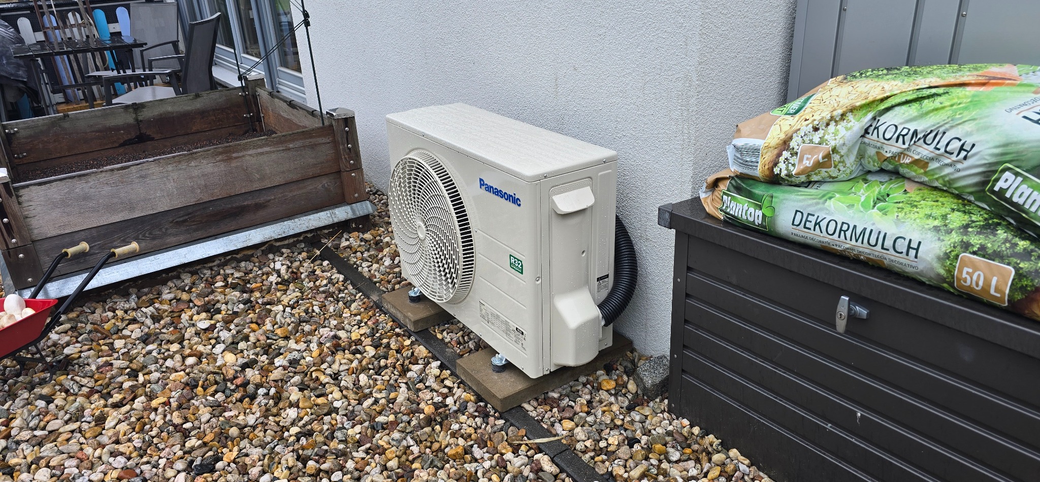 Zamontowana jednostka zewnętrzna klimatyzacji Panasonic na kamiennym podłożu, obok skrzyni ogrodowej i worków z korą dekoracyjną. Widok z boku.