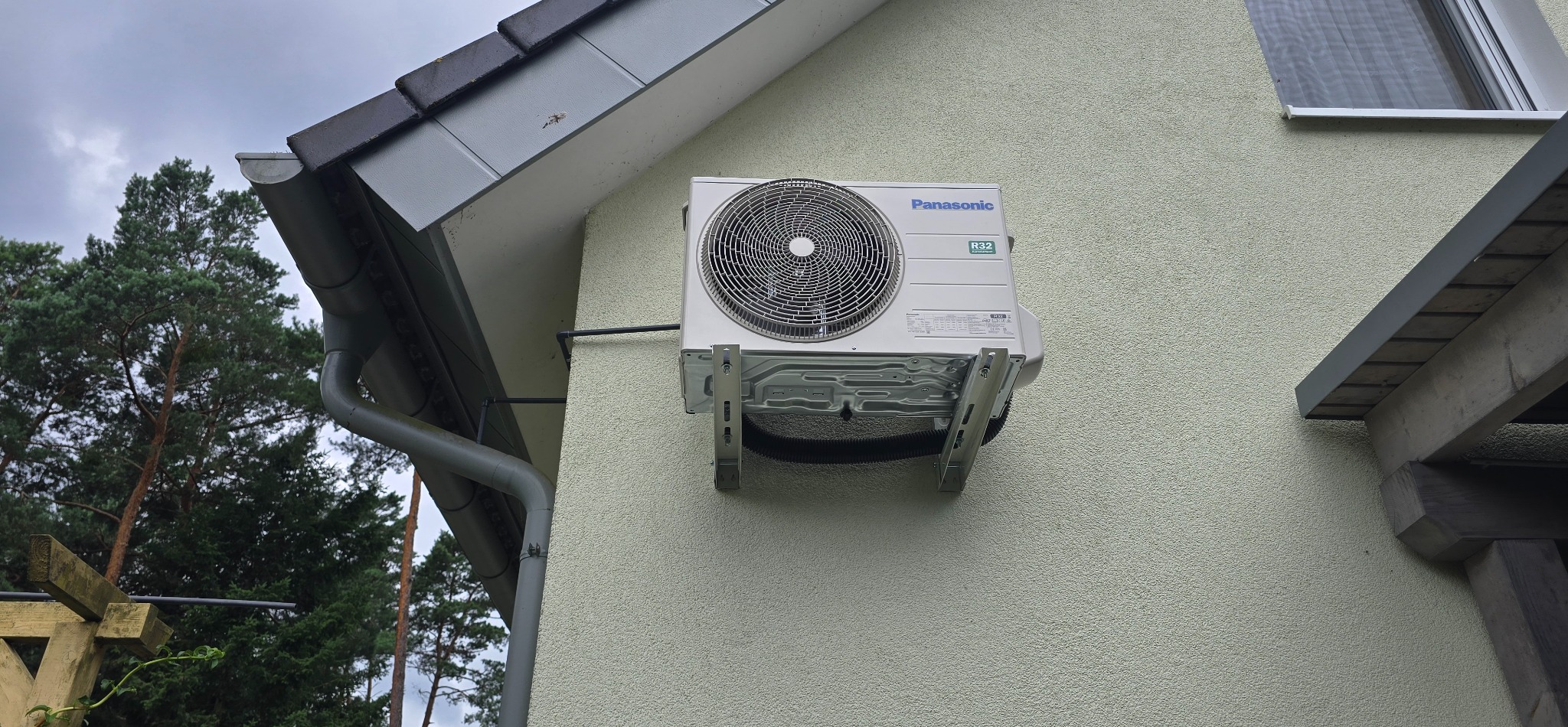 Zewnętrzna jednostka klimatyzacji Panasonic R32 zamontowana na elewacji domu z jasnym tynkiem, widoczny fragment dachu i rynny w szarym kolorze.