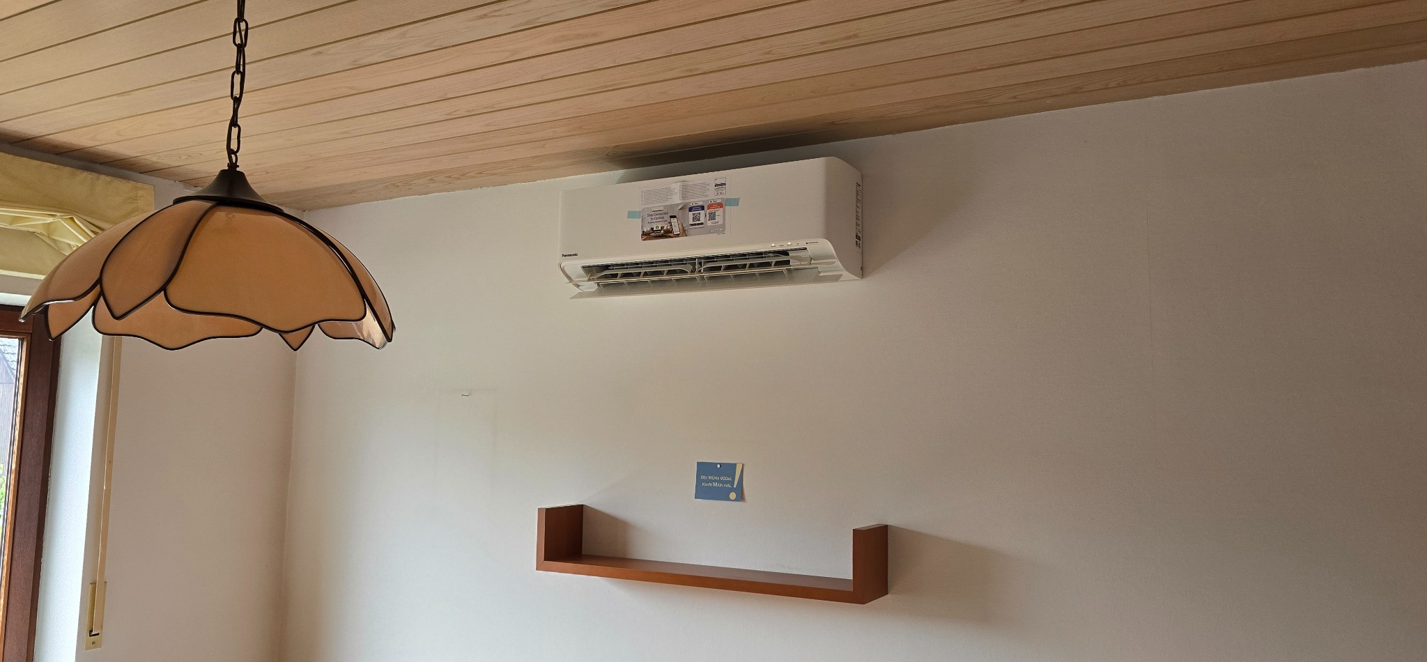 Biała klimatyzacja Panasonic zamontowana na ścianie w pokoju z drewnianym sufitem i lampą w stylu retro. Widoczna półka i karteczka z informacjami.