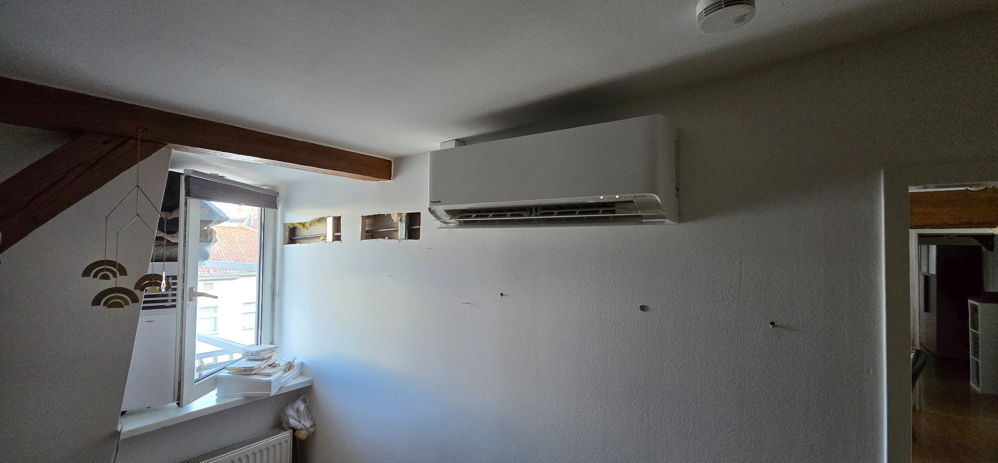 Biały klimatyzator na ścianie z widocznymi otworami po instalacji, w tle okno z dekoracją. Widoczne elementy drewniane konstrukcji dachu.