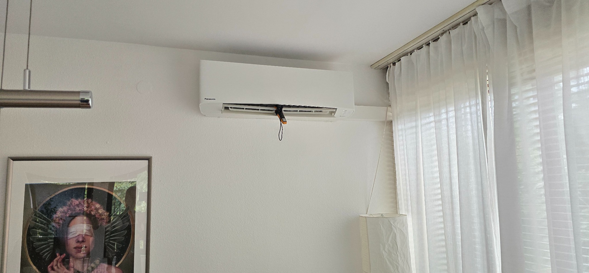 Biały klimatyzator Panasonic na ścianie w jasnym pokoju, z termometrem w kratce wentylacyjnej, sugerujący serwis. Obok okno z białymi zasłonami i lampa.