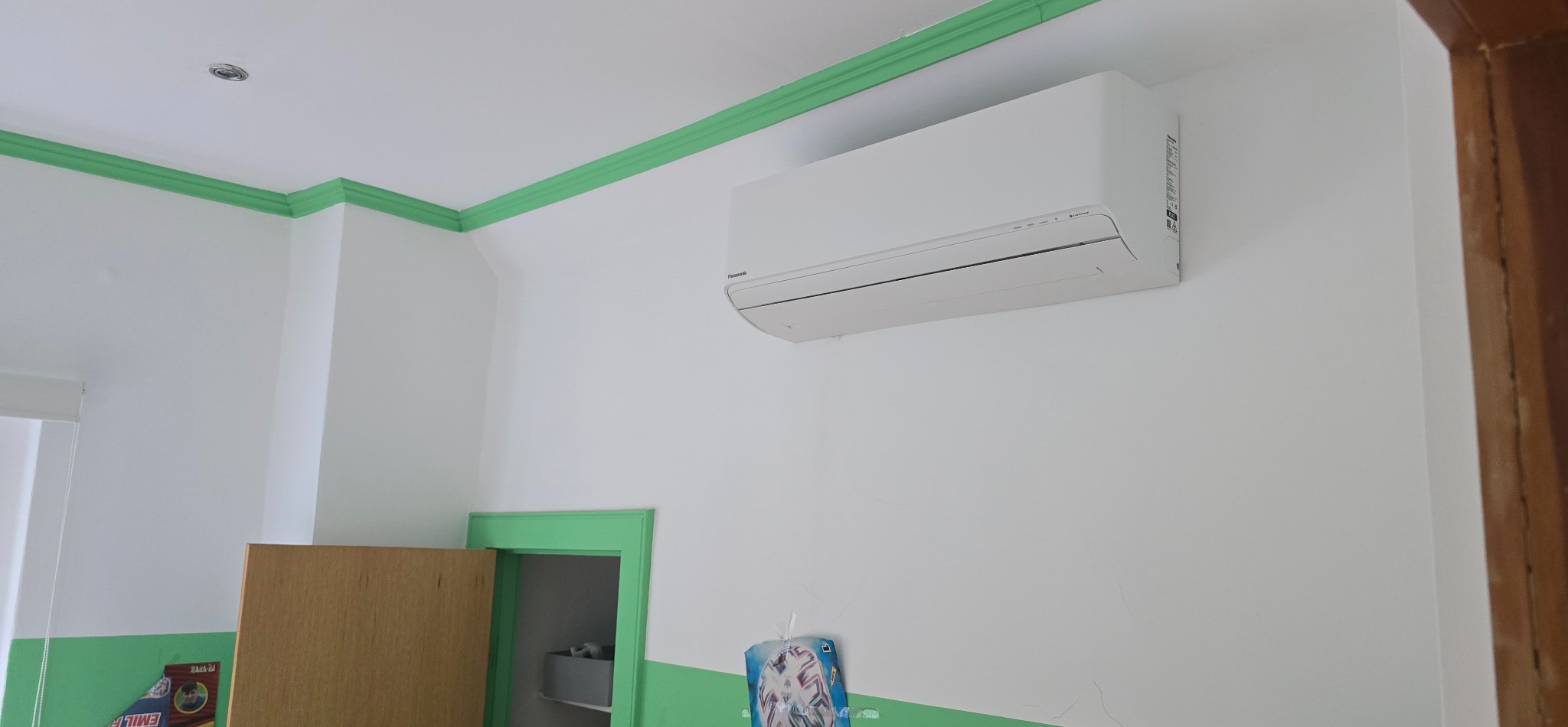 Biały klimatyzator Panasonic zamontowany na ścianie w pokoju z zielonymi listwami i futryną. Widoczny fragment szafki i drzwi.