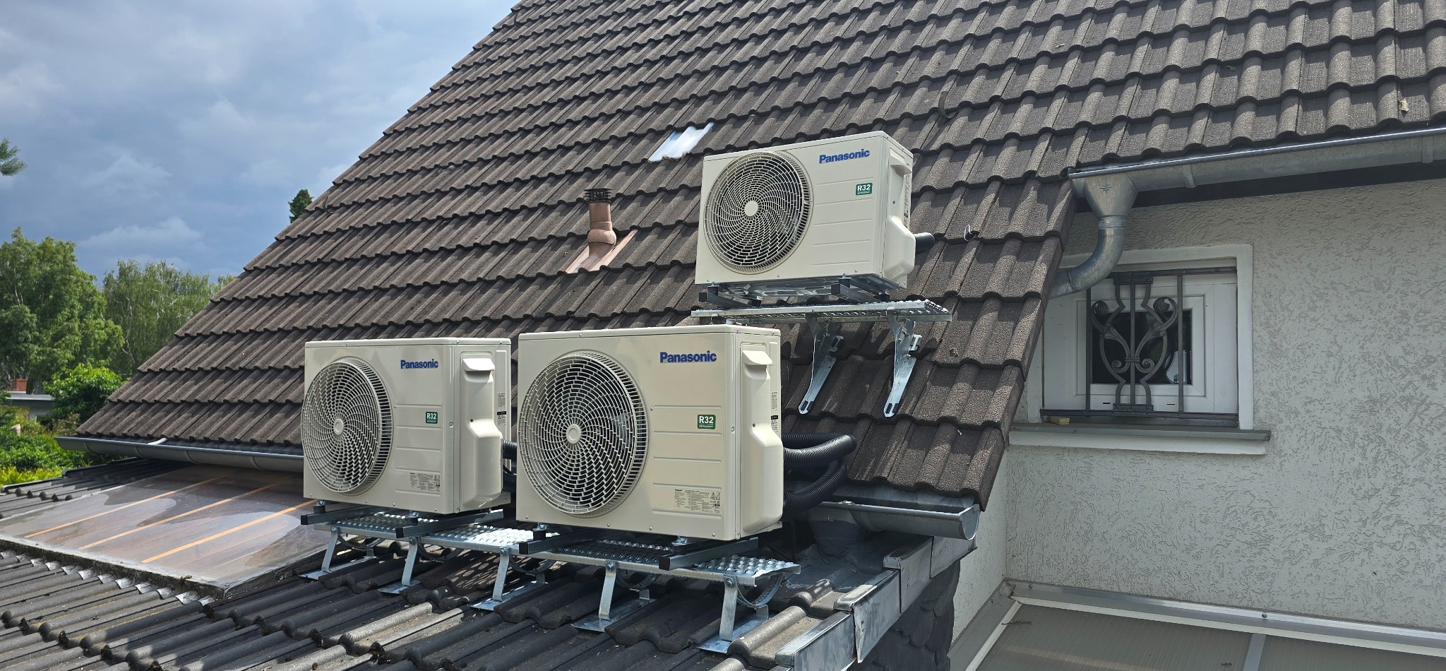 Trzy jednostki zewnętrzne klimatyzacji Panasonic zamontowane na dachu domu z brązową dachówką, na metalowej konstrukcji wsporczej, widok z dołu.