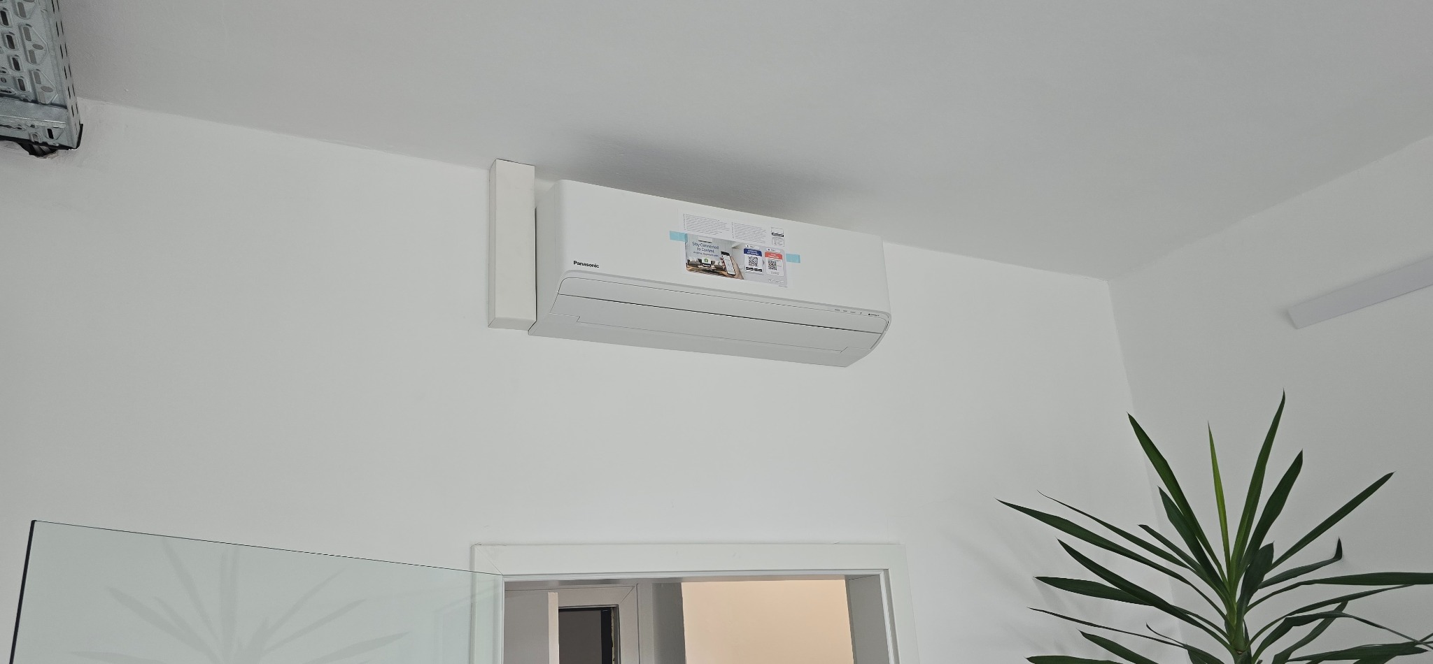 Biała klimatyzacja Panasonic na ścianie w jasnym wnętrzu, widoczna roślina doniczkowa i fragment szklanej balustrady. Minimalistyczny design.