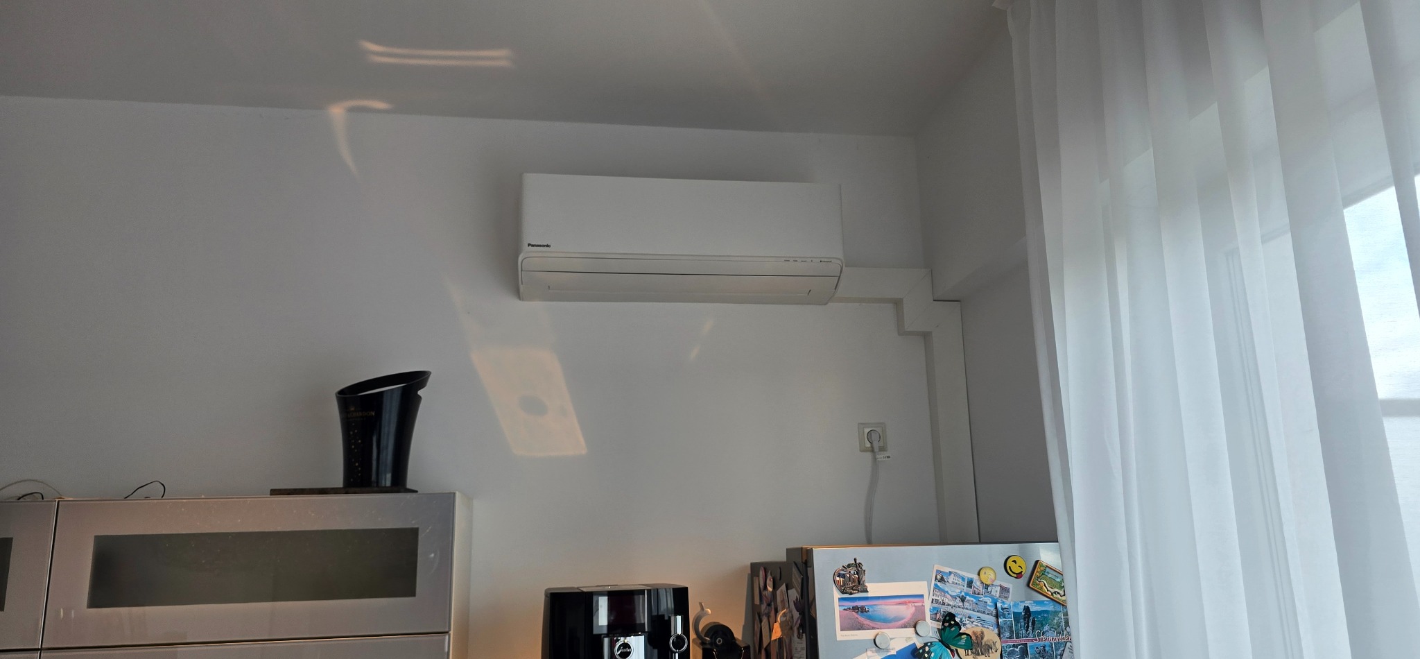 Biały klimatyzator Panasonic zamontowany na ścianie w jasnym wnętrzu, widoczne elementy wyposażenia kuchni i okno z firanką.